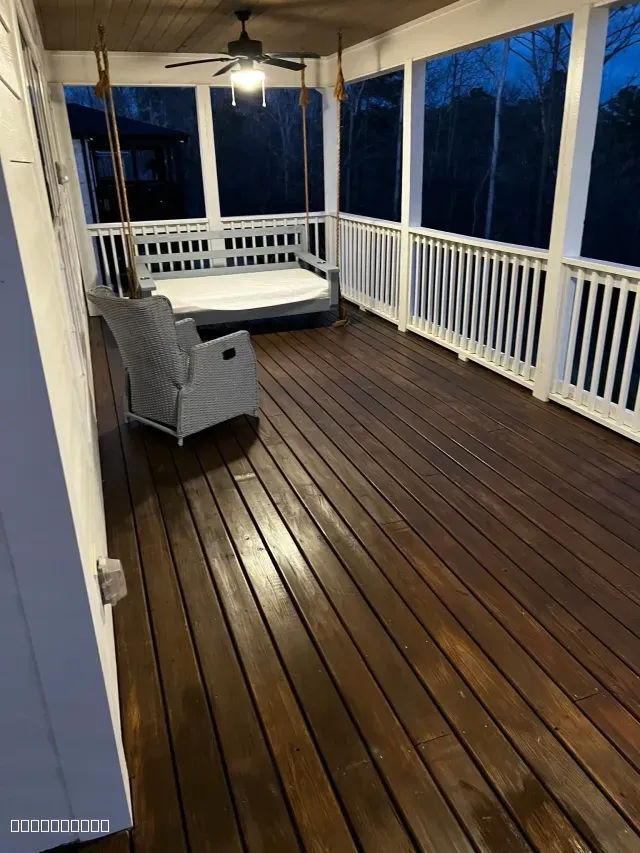 A covered porch with freshly stained dark wood floors, white railings, a hanging swing, and a wicker chair.