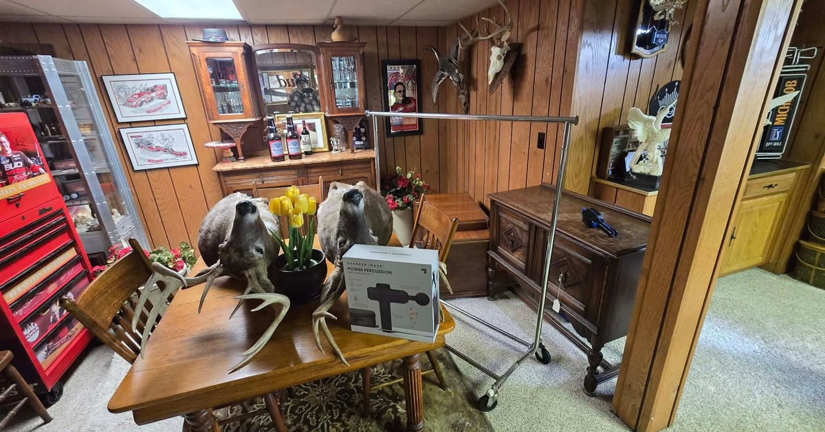 Rustic wood-paneled basement man cave featuring deer head mounts, bar area with liquor shelves, central table with floral decor and cross.