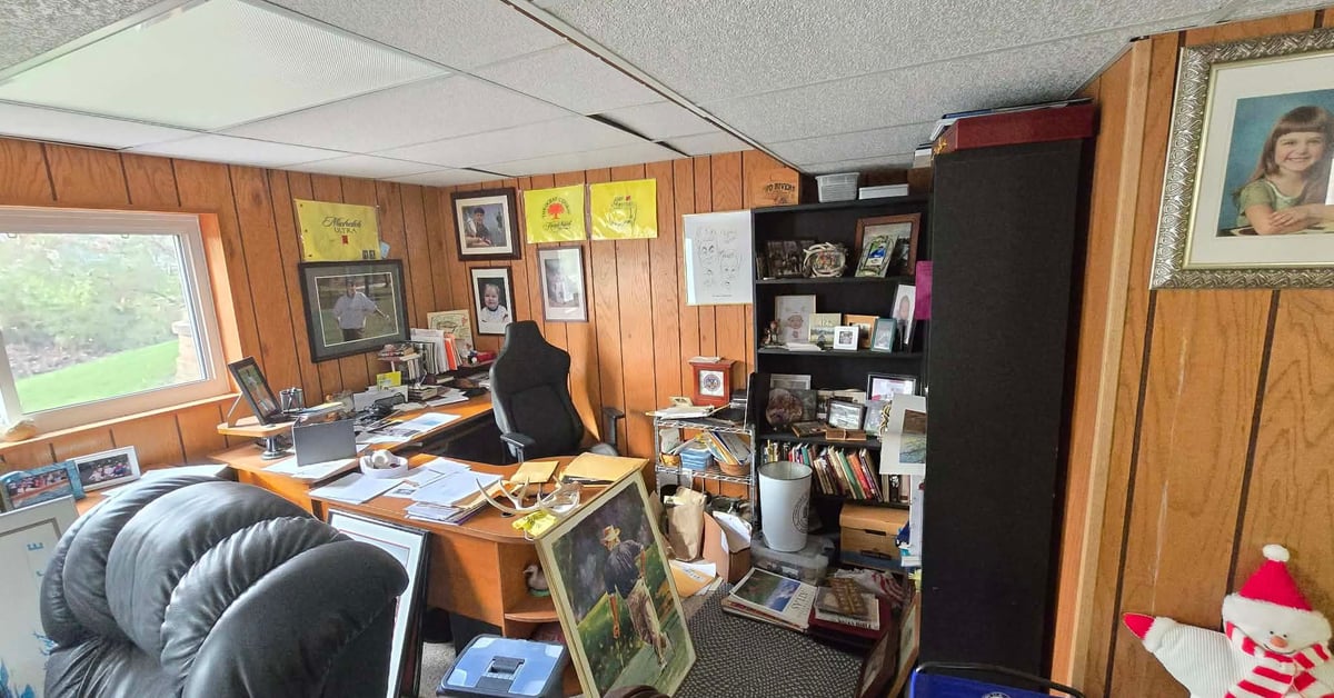 Cluttered basement home office with wood-paneled walls, desk with monitors, bookshelves, chairs, and decorations.