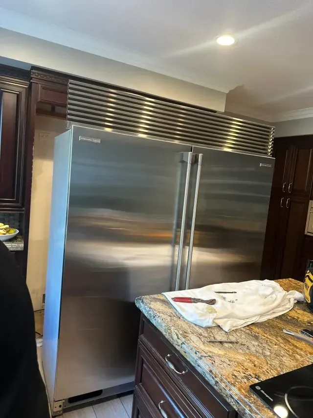 A stainless steel built-in refrigerator being serviced in a kitchen with dark wood cabinets and granite countertops.