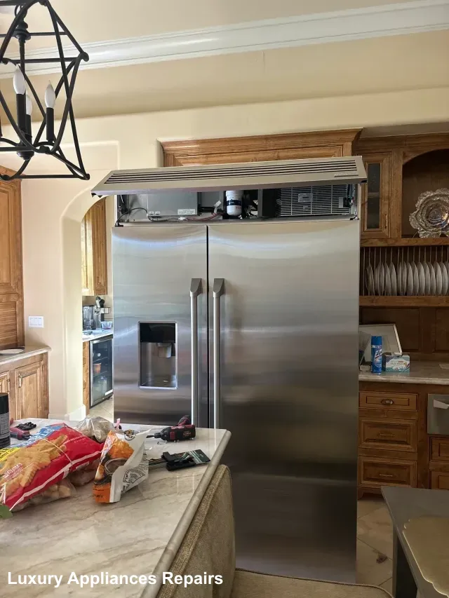A stainless steel built-in refrigerator with the top service panel open for repair in a kitchen.