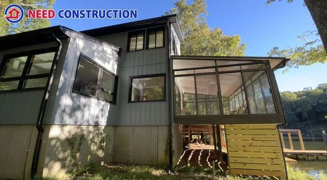 A modern two-story home with gray siding and a large glass sunroom addition overlooking a lake.