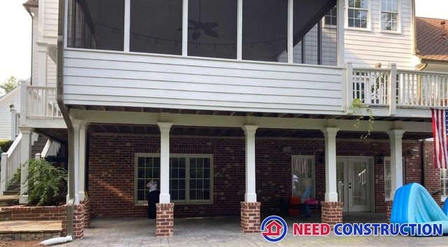 A two-story outdoor living space featuring a white screened-in porch above a paved patio with an underdeck drainage system.