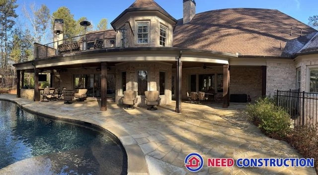 A luxurious custom outdoor living space featuring a stone patio, swimming pool, and covered lounge area by Need Construction.