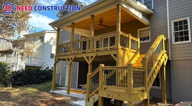 A large two-story wooden deck and covered porch with a ceiling fan and black railings attached to a gray house.