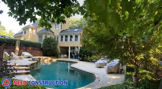 A large backyard with a custom swimming pool, stone patio, and a sunroom home addition under a canopy of green trees.