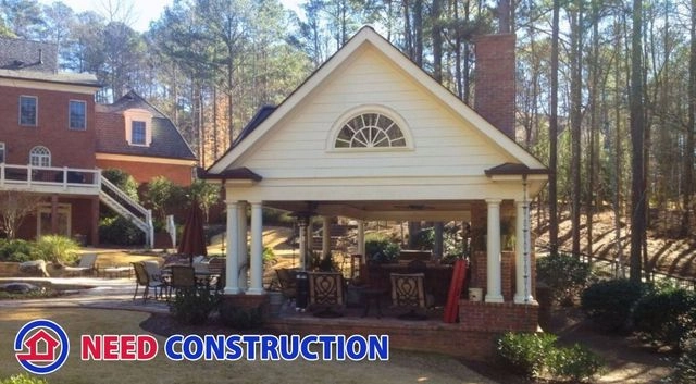 A luxurious outdoor living pavilion with a brick fireplace and white columns in a wooded backyard.