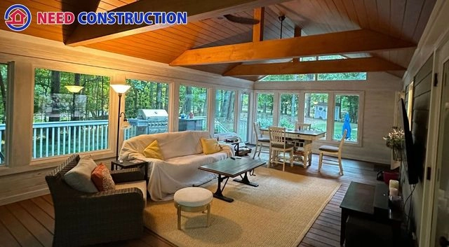 A luxurious sunroom with large windows, vaulted wood ceiling, and comfortable living room furniture overlooking a forest.
