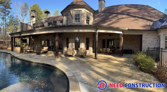 A luxurious backyard with a stone patio, swimming pool, and covered outdoor living space by Need Construction.