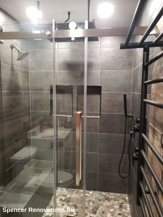 A newly remodeled modern bathroom shower featuring dark gray wall tiles, a glass enclosure, a pebble stone floor, and a black towel warmer.