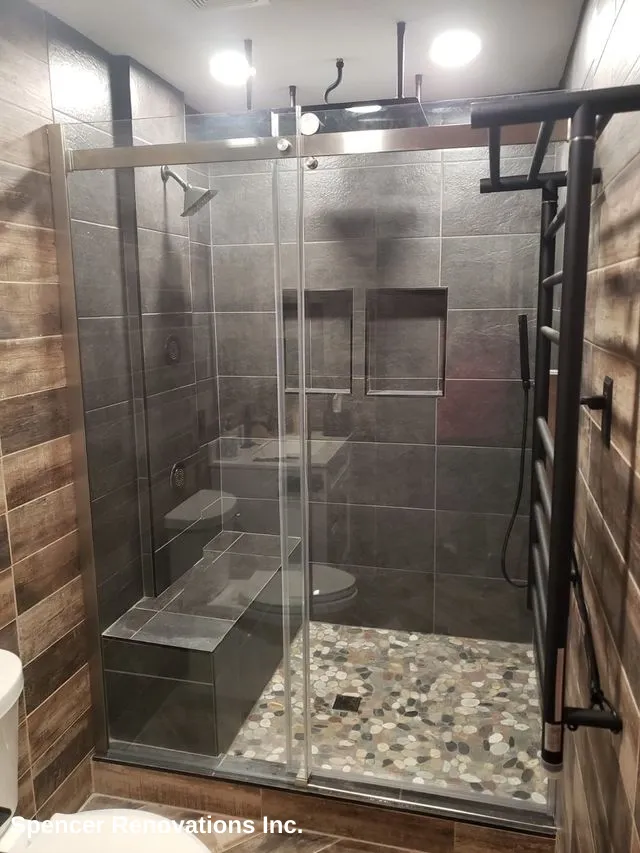 A modern walk-in shower with dark grey tiles, a glass enclosure, built-in bench, and a pebble-stone floor, alongside a black towel warmer.