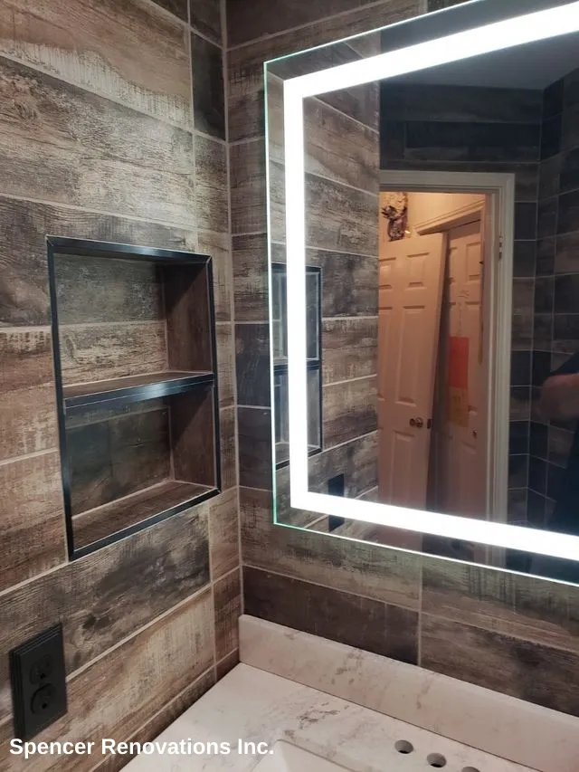 A modern bathroom after renovation, featuring wood-look tile walls, an LED-lighted mirror, a built-in shower niche, and a white vanity with a sink.