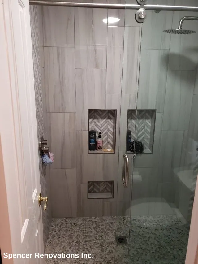 Modern bathroom shower with light grey wall tiles, pebble floor, glass enclosure, and decorative storage niches.