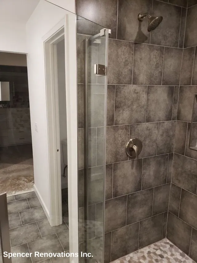 A modern, newly remodeled bathroom shower featuring dark gray rectangular wall tiles, a pebble-tiled floor, and a clear glass door with brushed nickel fixtures. A glimpse of the rest of the bathroom with gray floor tiles and a vanity is visible through the doorway.