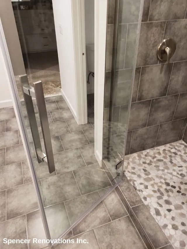 A modern walk-in shower with glass doors, dark grey tiled walls, and a light pebble-stone shower floor in a newly remodeled bathroom.