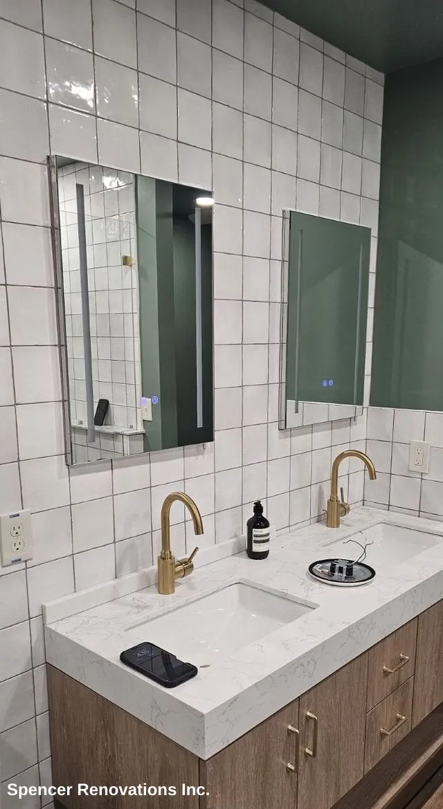 A modern white freestanding bathtub with a wall-mounted brass faucet, set against white square tiles and a dark green wall, with white hexagonal mosaic floor tiles.