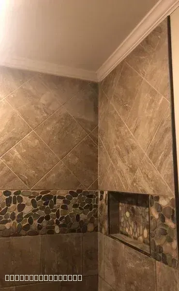 Professional tile installation in a shower featuring diagonal stone tiles and a pebble accent strip with a recessed niche.