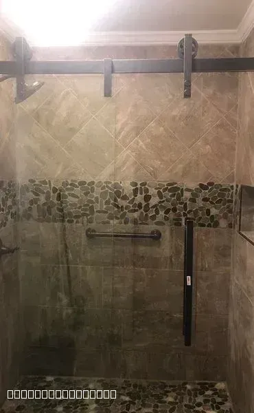A modern walk-in shower featuring tan wall tiles, a pebble accent strip, and industrial sliding glass door hardware.