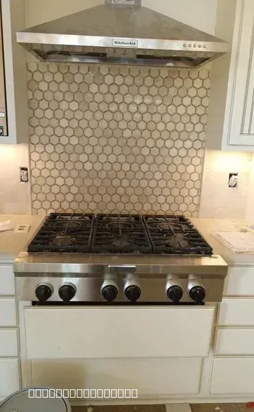 A close-up of a kitchen featuring a new hexagon tile backsplash behind a stainless steel stove and under a range hood.