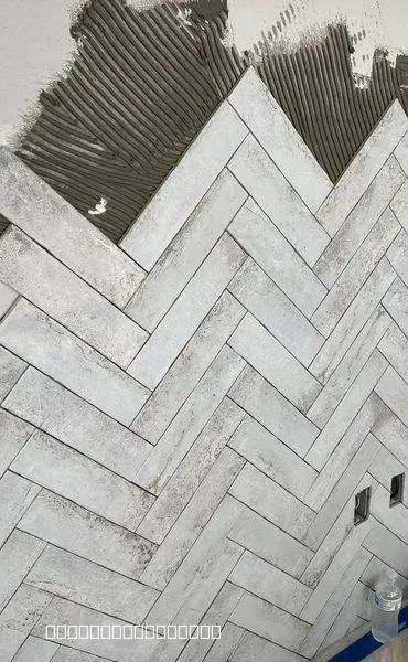 Close-up of light gray tiles being installed in a herringbone pattern on a wall with visible mortar.